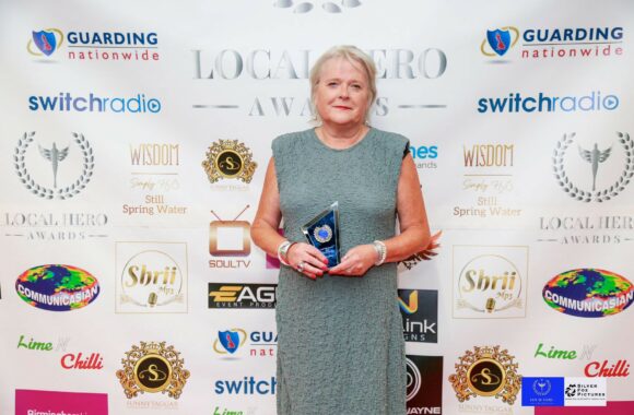 Domestic abuse support worker Ruth Carton holds her award in front of a media board while wearing a cocktail dress.